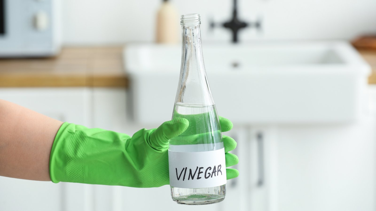 A gloved hand holding a clear bottle labeled "vinegar" in a kitchen setting.