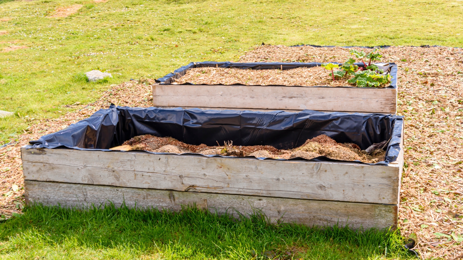 A newly constructed wooden rectangular structure filled with dark soil. There is a black plastic liner partially covering the soil. The surrounding ground is a mix of short, green grass and some bare patches.