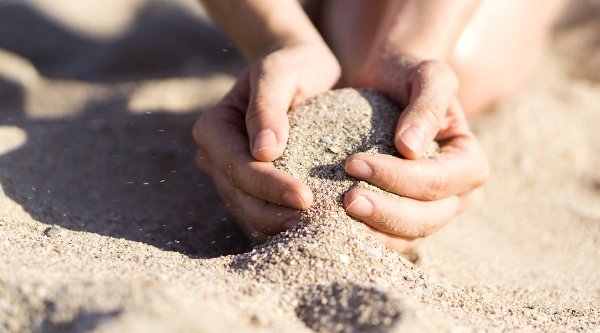 Close-up of women's hands picking up sand in their palms. The sand appears as a vast expansion of fine grains. The texture is soft and smooth, with occasional ripples and patterns shaped by the wind. The color is delicate golden beige.